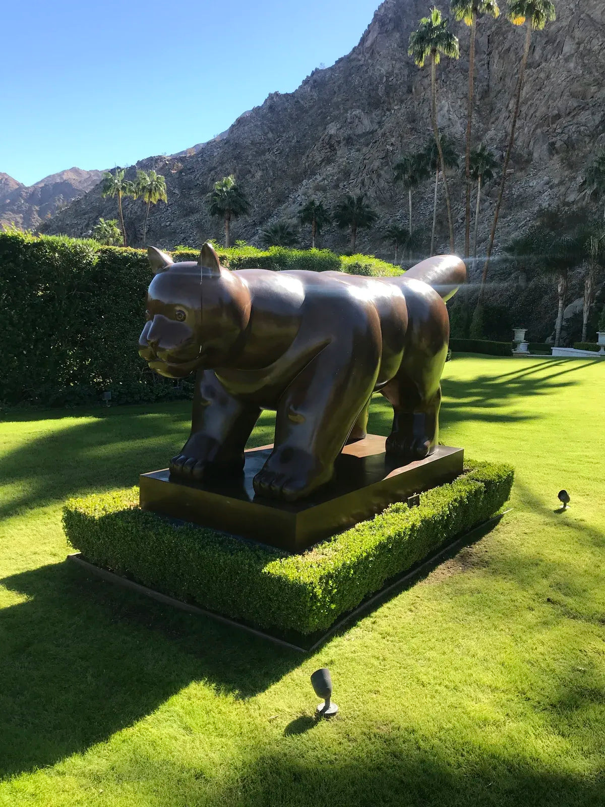 Large bronze big cat sculpture on a garden pedestal, with palm trees and mountains in the background.