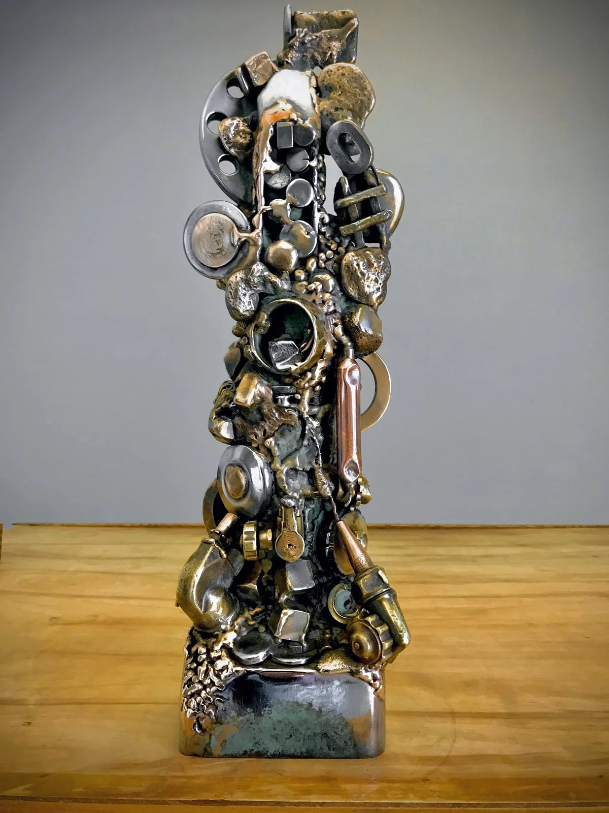 An abstract metal sculpture made from stacked and welded industrial parts, including gears, pipes, and circular metal elements, displayed on a wooden surface against a neutral gray background.