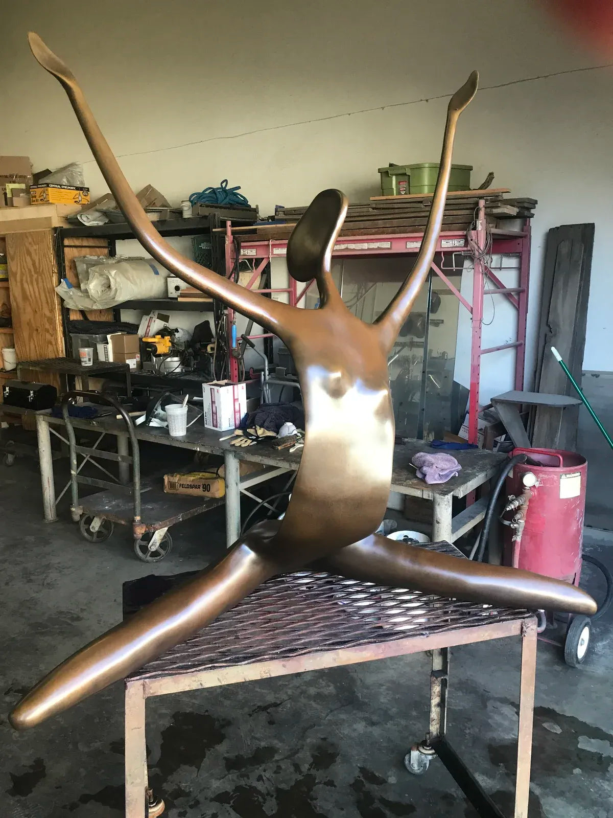A bronze-finished abstract sculpture of a stylized human figure with outstretched arms and legs, displayed on a metal workbench in a cluttered workshop.