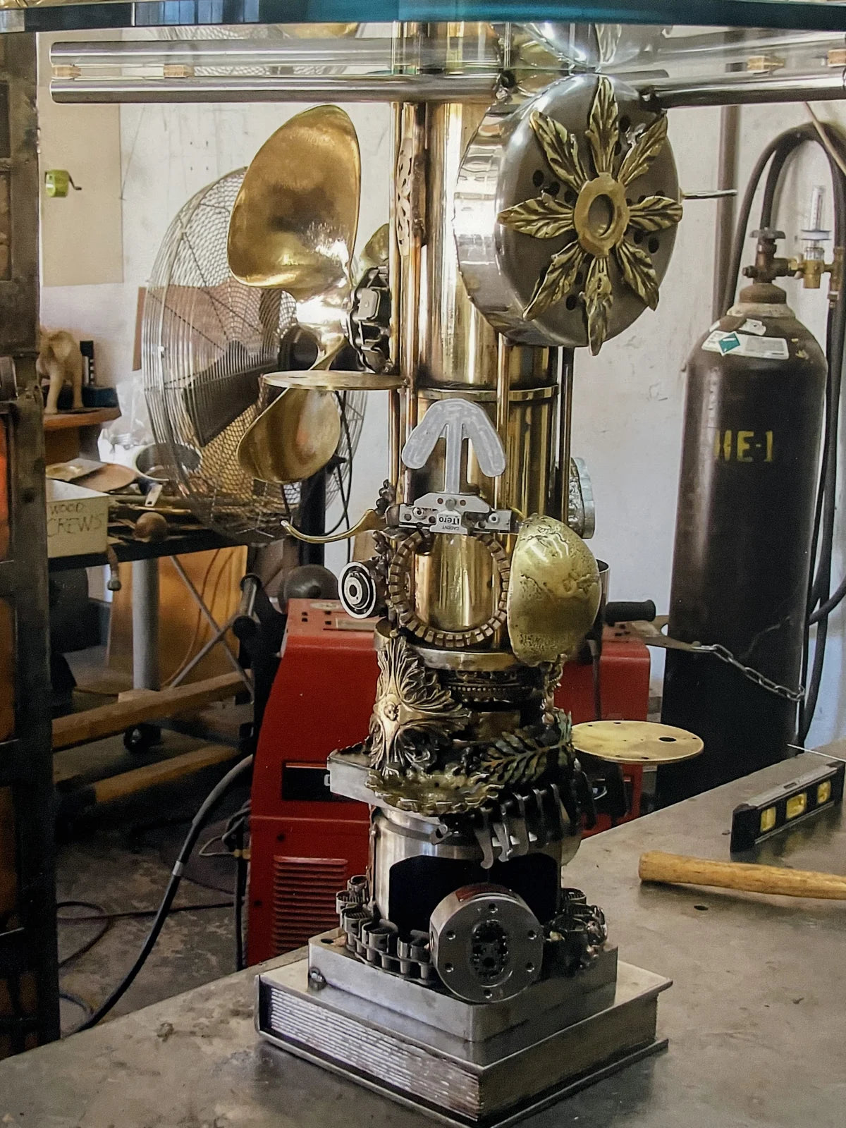 A steampunk-style decorative metal sculpture or machine centerpiece with brass and silver metallic components, gears, and ornate detailing, displayed on a workbench in a workshop with tools and equipment visible in the background.