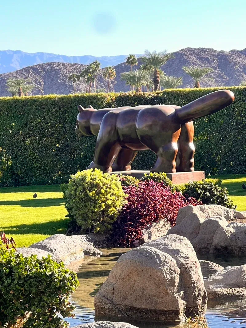 A large metallic cat sculpture standing on a base in a landscaped garden with rocks, shrubs, grass, hedges, palm trees, and mountains in the background.