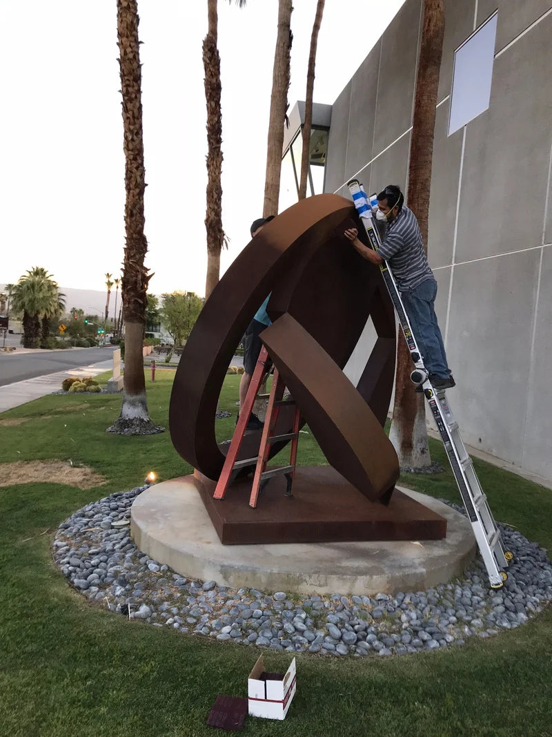 A large abstract metal sculpture in a circular interlocking design is being installed outdoors on a circular concrete base. Two workers are visible on ladders, adjusting the sculpture near palm trees and a modern building wall.