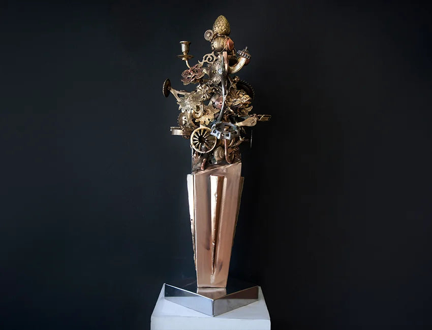 A steampunk-style sculpture featuring intricate metallic gears, wheels, and mechanical details in gold and bronze tones, mounted on a tall reflective metallic pedestal and displayed on a white geometric base against a dark background.