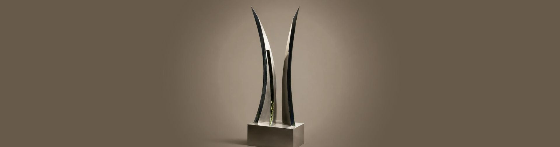 A modern abstract metal sculpture with two tall, curved, blade-like forms rising from a rectangular base, shown against a plain warm brown studio background.